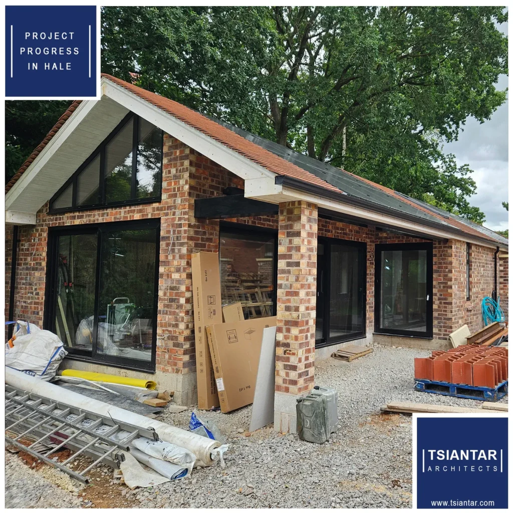 Partially completed brick house with large glass windows under construction, surrounded by building materials. Signs read "Project progress in Hale" and "Tsiantar Architects.